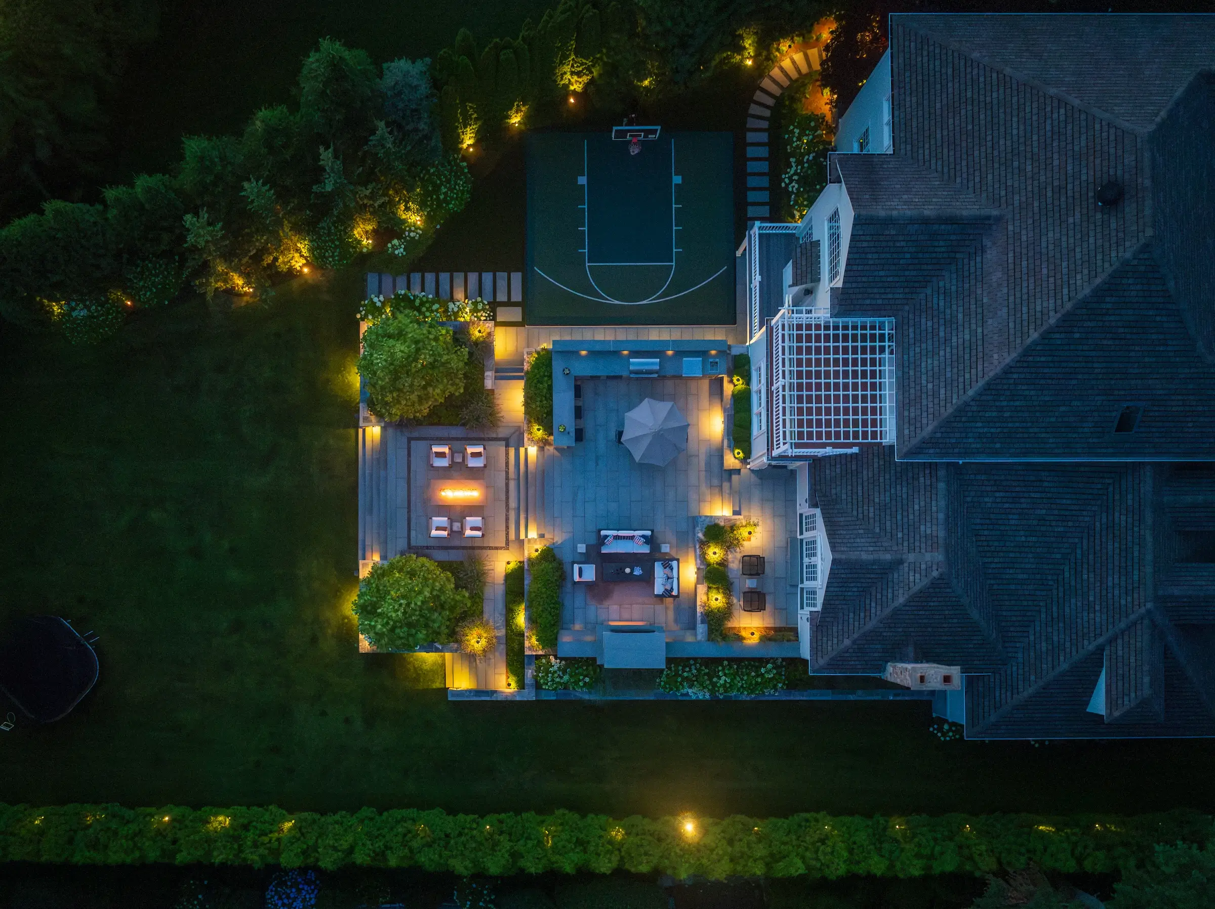 An aerial bird's-eye view of a Sudbury, MA estate at twilight. Warm landscape lighting illuminates a multi-level stone patio with a lounge area, outdoor kitchen, and fire pit. Next to the patio is a custom dark green basketball half-court, all surrounded by a manicured lawn and privacy hedges.