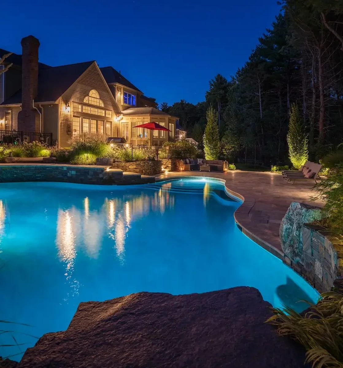 Night view of a lit, curved swimming pool in a luxury backyard with a illuminated house and outdoor seating area.