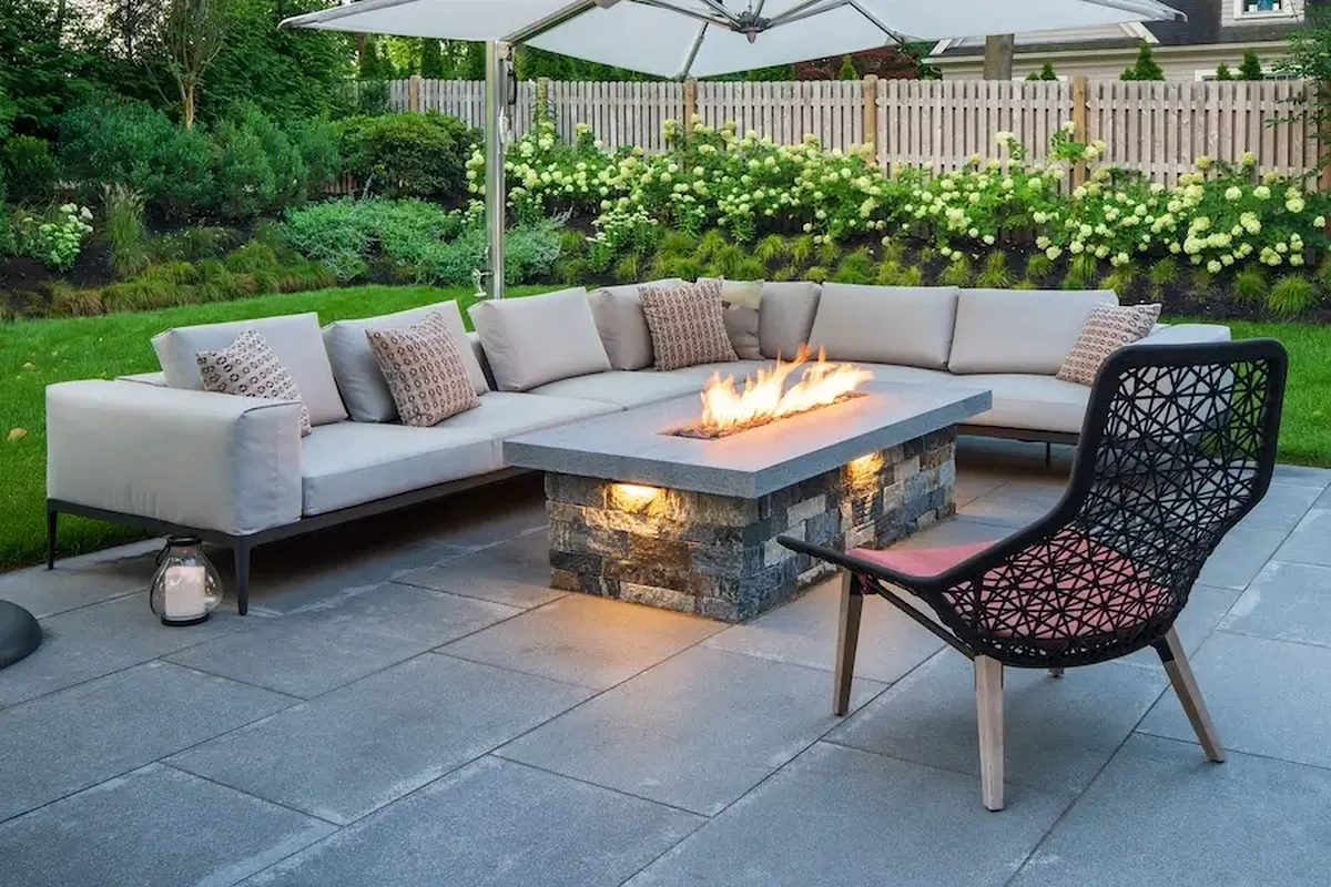 Outdoor patio with a gray sectional sofa surrounding a rectangular stone fire pit under a large umbrella while a woven black chair sits nearby.